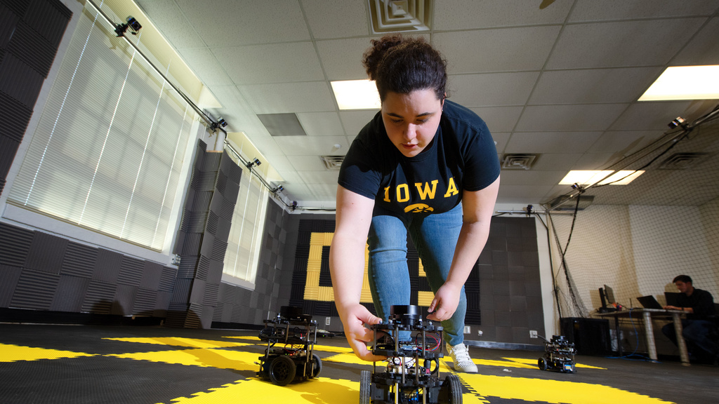 Camilla Tabasso, Undergraduate Student, bends over facing the camera to pick up a small robot car
